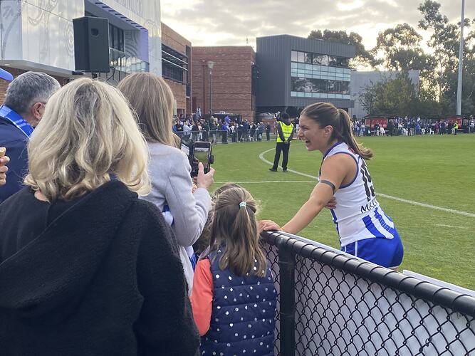 Spectators seen from behind football oval fence interacting with smiling female footballer on standing on oval.