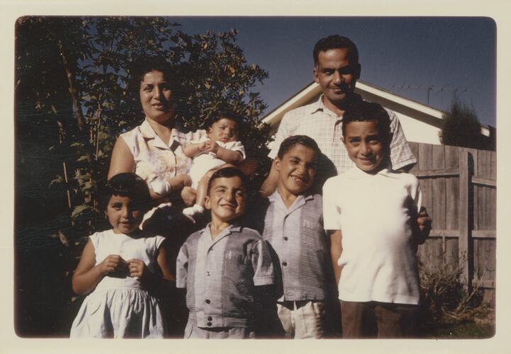 Woman and man posing outside with 5 children. The woman holds a baby. Garden, fence and white weatherboard behind them.