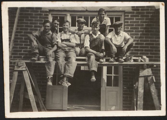 Man sitting on a trestle with six workmates on a building site. Wooden doorway behind them has sidelight and transom windows. The building is made of brick.