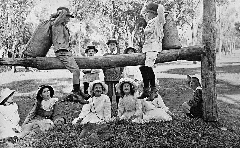 Two small boys, straddling a log, fighting with hay filled sacks. There is hay spread on the ground to break their fall and they are watched by ten other children.