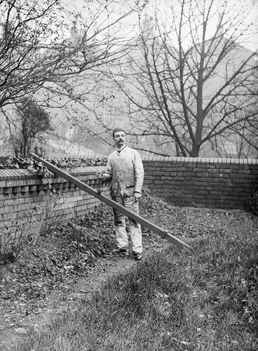 Man stands in a walled garden wearing white working clothes. He's behind a pipe propped against the wall.
