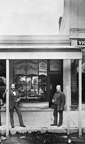 Two men standing on footpath, under a verandah, in front of a tobacconist and wine Merchant shop.