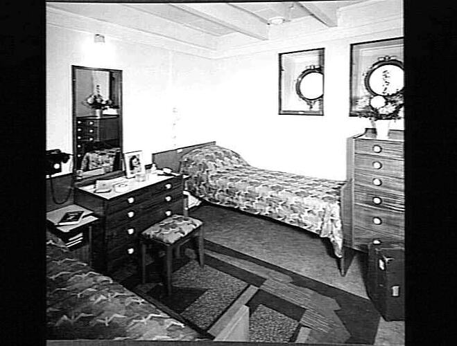 Ship interior. Two single beds, one against wall. Dressing table and stool in between. Drawers at right.