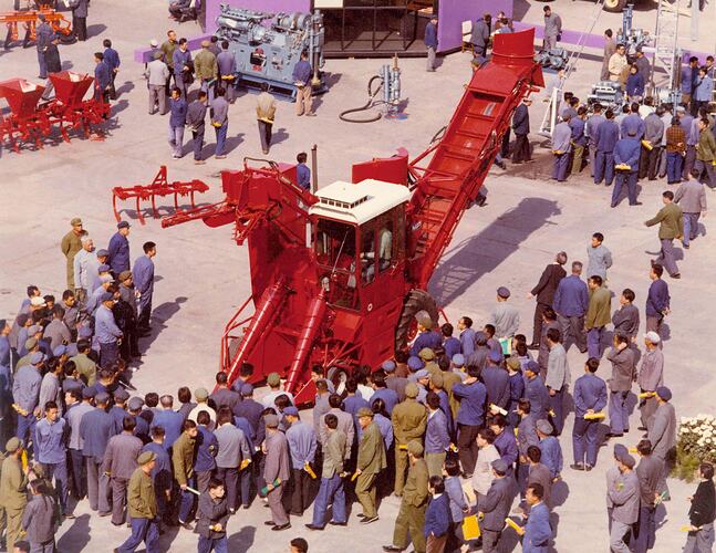 Large group of people stand around a cane harvester at an exhibition.