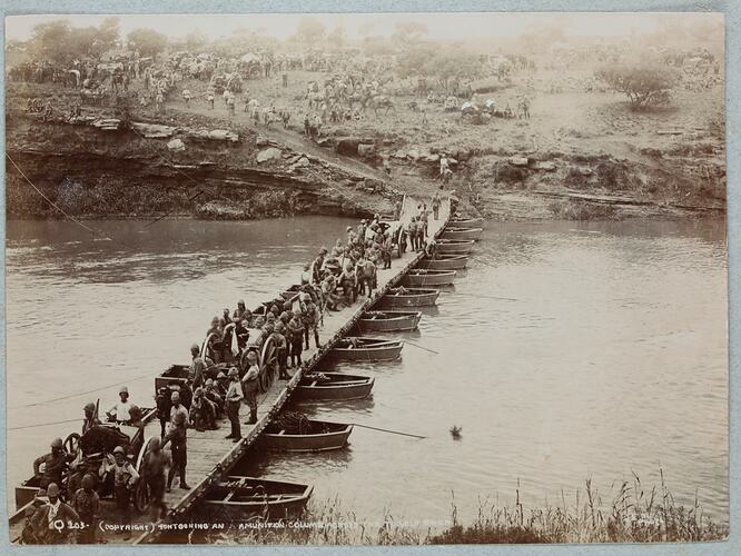 Boats supporting floating wooden bridge with large group of soldiers on bridge and on land nearby.