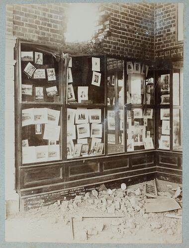 Display case containing photographs with hole in wall above case and rubble on ground.